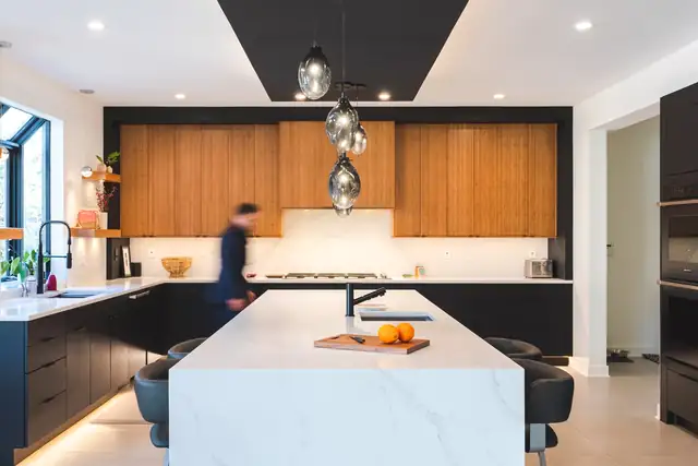 Potomac, MD kitchen with wood-grain upper cabinets set into a black wall, black ceiling soffit, smoked glass pendants and a white waterfall island