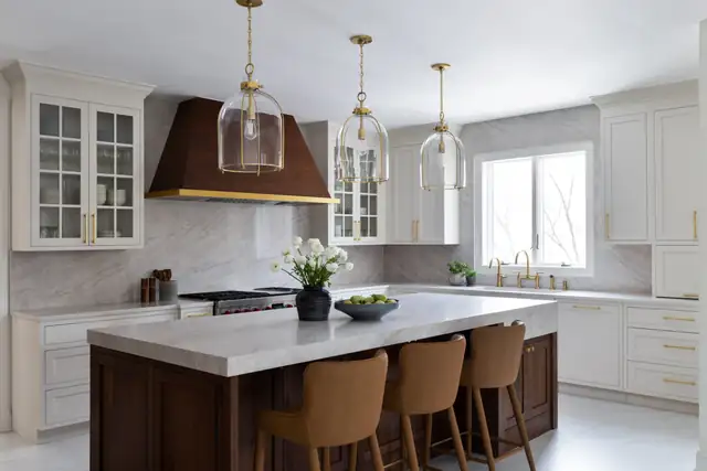 White shaker kitchen with a walnut and brass range hood, a dark wood island with stone top and clear glass pendants